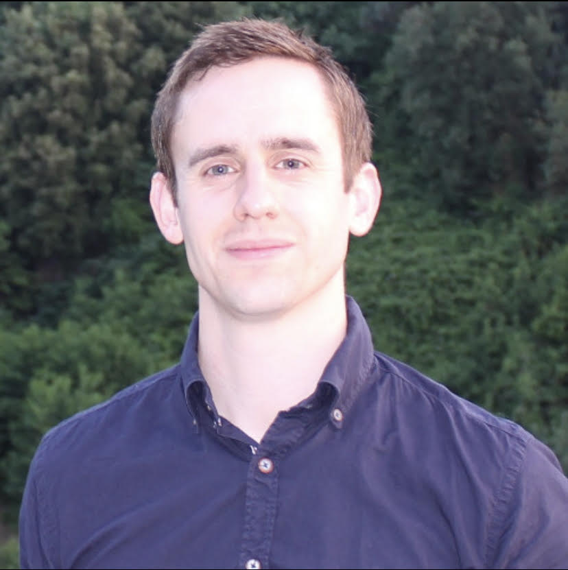 A photograph of an employee of the Gambling Commission looking towards the camara. The employee is wearing a purple shirt and is standing in front of trees which can be seen in the distance.  