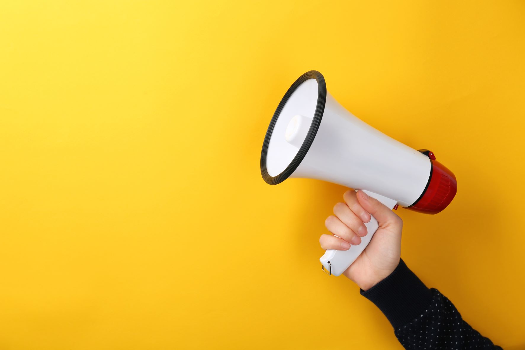 A yellow background showing a person's right-hand raised holding a microphone to the sky. The person is wearing a black woolly jumper. The microphone is white with a red back section.