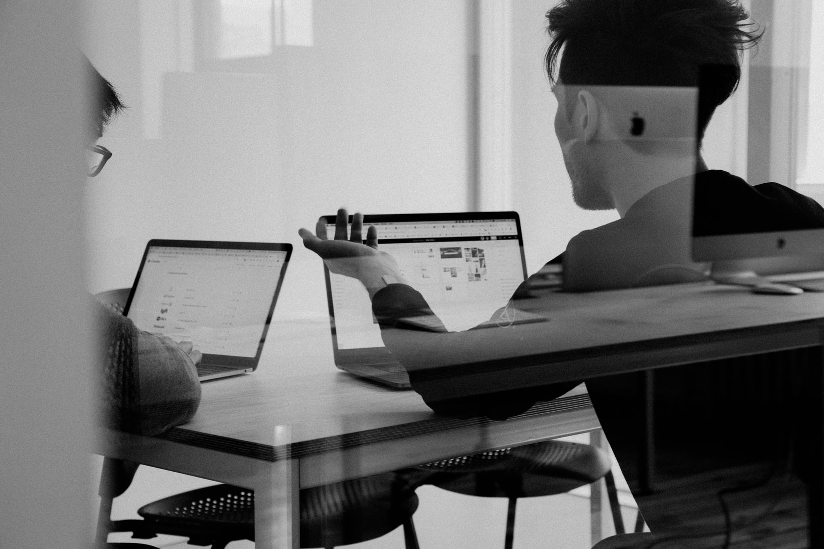 A photograph, taken through some glass, of two individuals sitting at a table with laptops in front of them. The individuals have their back to the camara. The laptops appear to be displaying spreadsheets and graphs. One individual has their arm raised and appears to be discussing what is on their screen to the other individual.